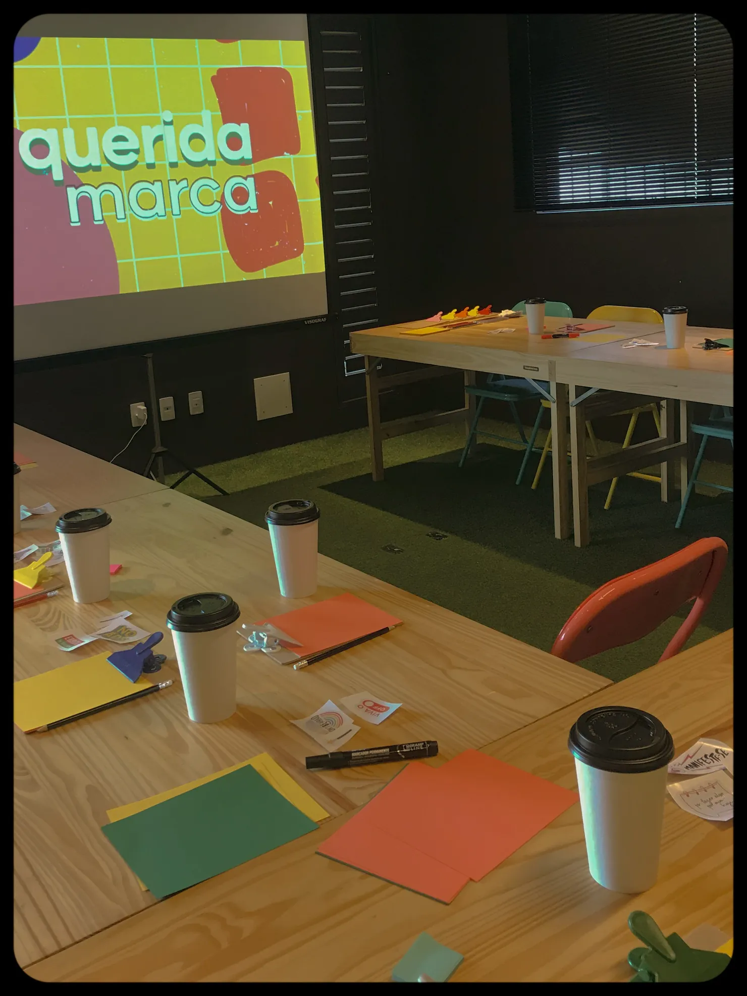 Sala de workshop com "querida marca" projetado na parede e papéis coloridos sobre as mesas