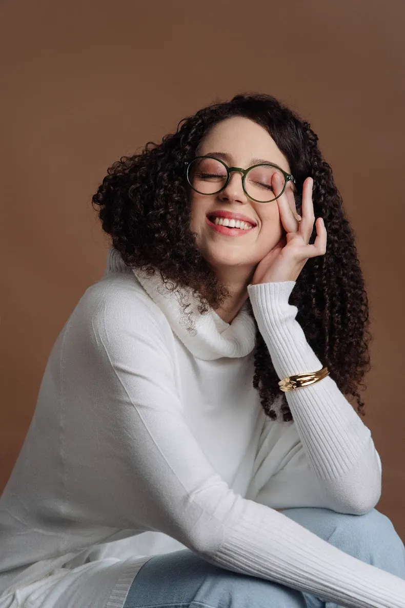 Mulher de óculos, sorrindo de olhos fechados, com a mão no rosto